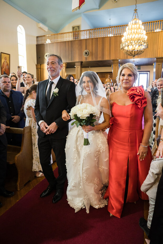 Bride in a lace wedding gown with veil and white bouquet escorted down the aisle by her father in a black suit, with a woman in a red dress beside them in a church.