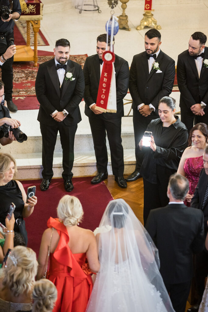 Bride in a white gown and veil walks toward the altar as four groomsmen in black tuxedos stand along the aisle; guests take photos.