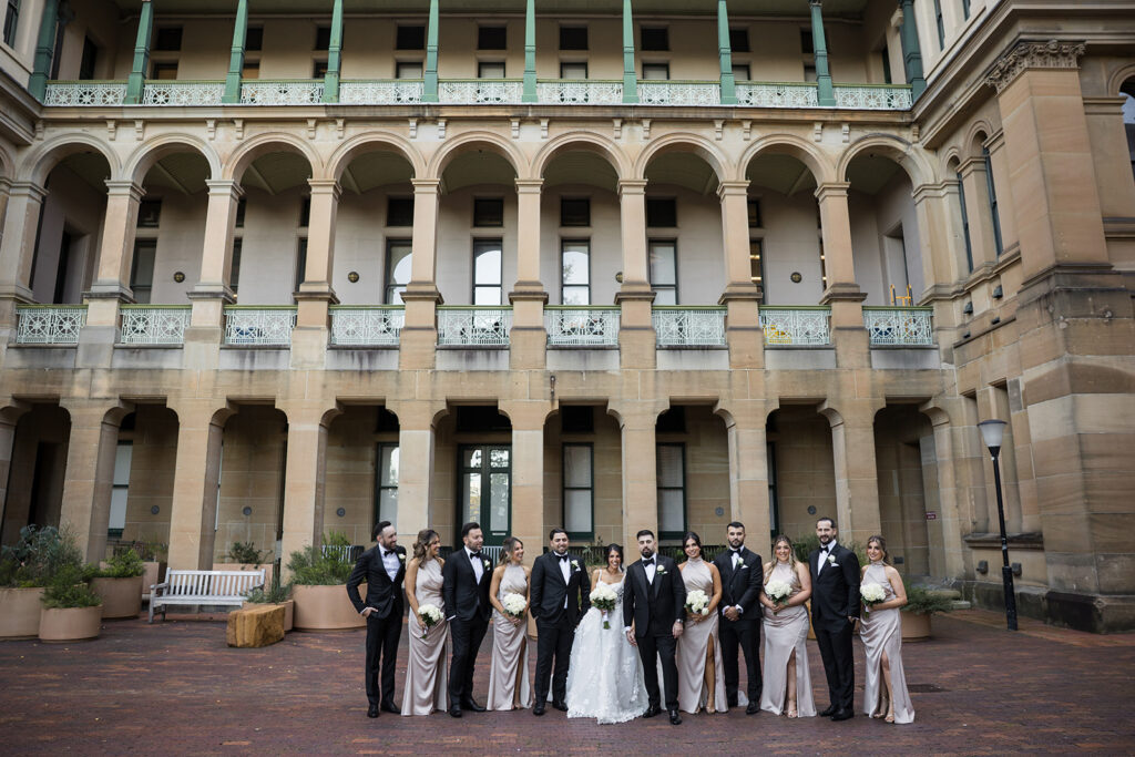Wedding party posing in front of a historic beige building with arched columns; brides in white gown, bridesmaids in champagne dresses, grooms in black tuxedos, holding bouquets.