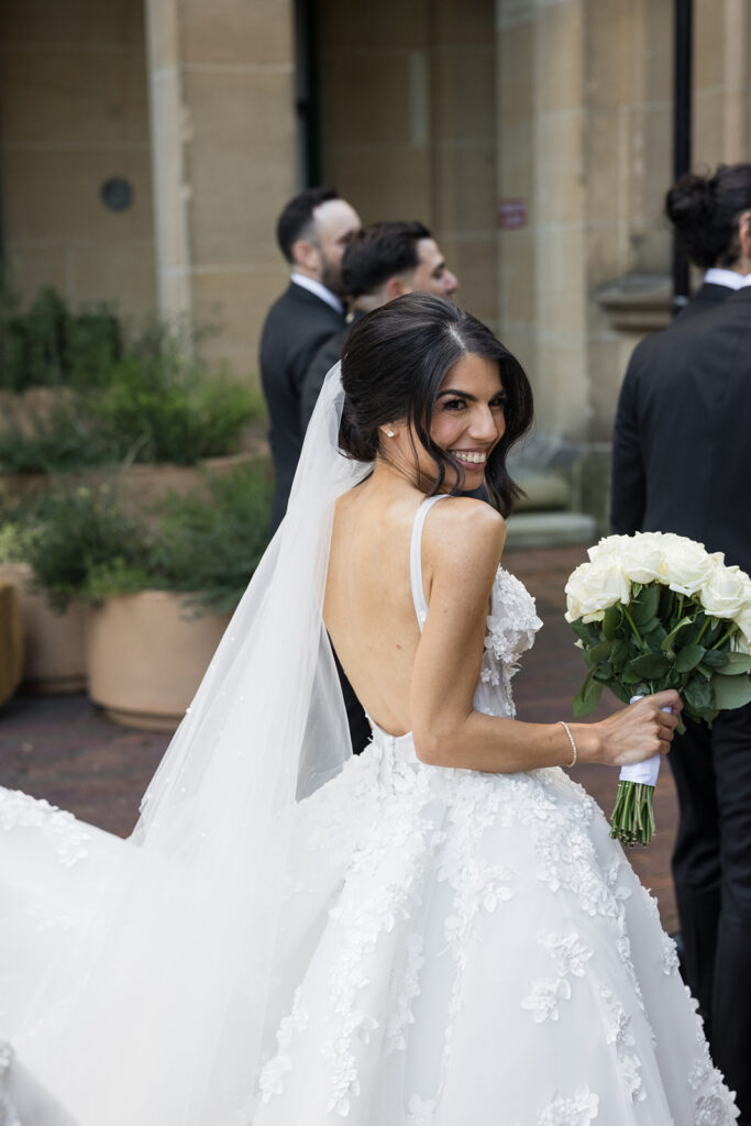 Bride in a white lace wedding dress turns and smiles over her shoulder, holding a bouquet of white roses as her veil trails behind her, with groomsmen in black suits in the background outside a stone building.