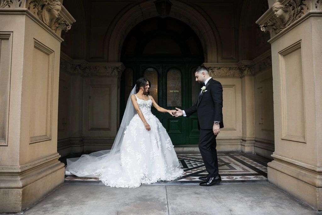 Bride in a lace wedding gown and long veil, extending a ring to a groom in a black suit under an ornate archway.