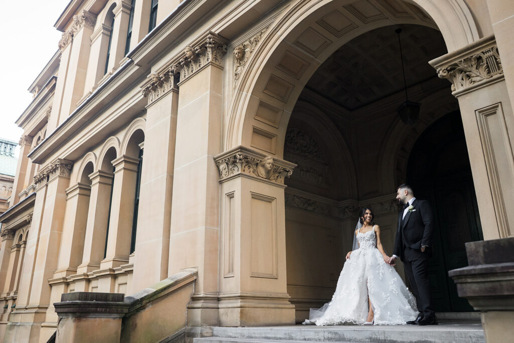 Bride in a white lace gown and veil, and a groom in a black suit, stand on stone steps under an ornate arched entrance of a historic beige building.