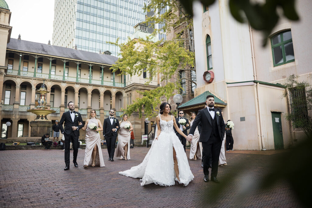 Bride in a white wedding gown walks with her wedding party in a historic courtyard, bridesmaids in beige dresses and groomsmen in black tuxedos.