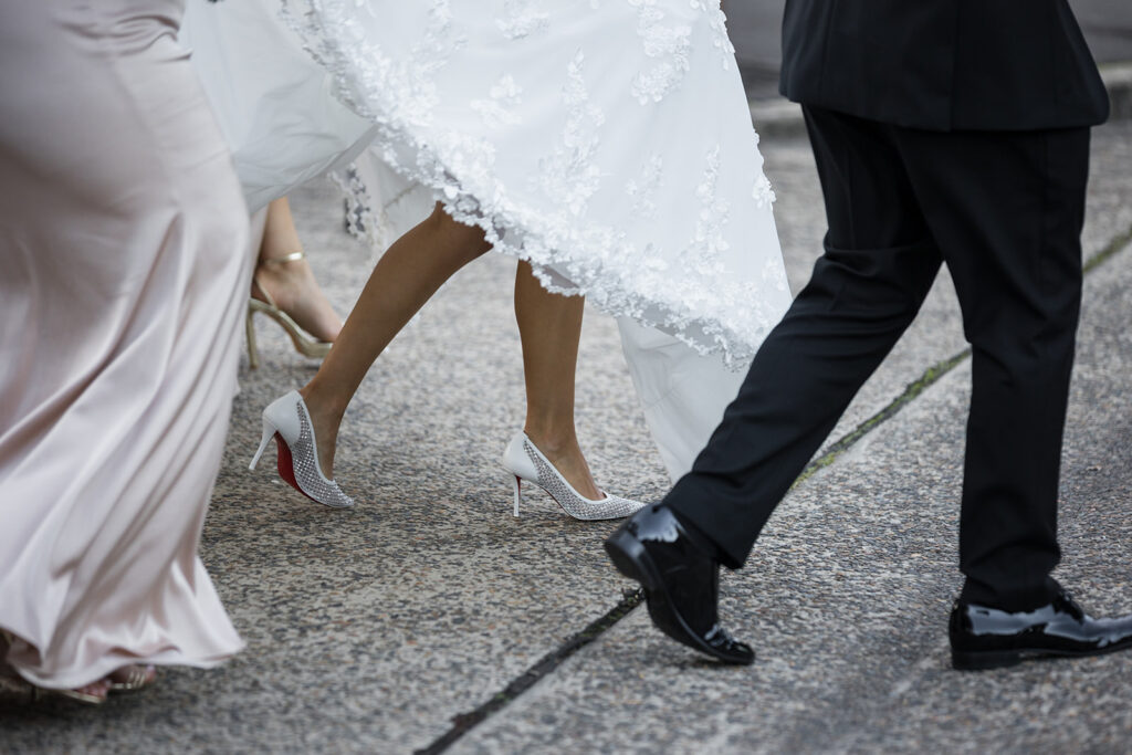 Bride in a lace wedding gown steps forward in glittery silver heels as guests walk beside her.