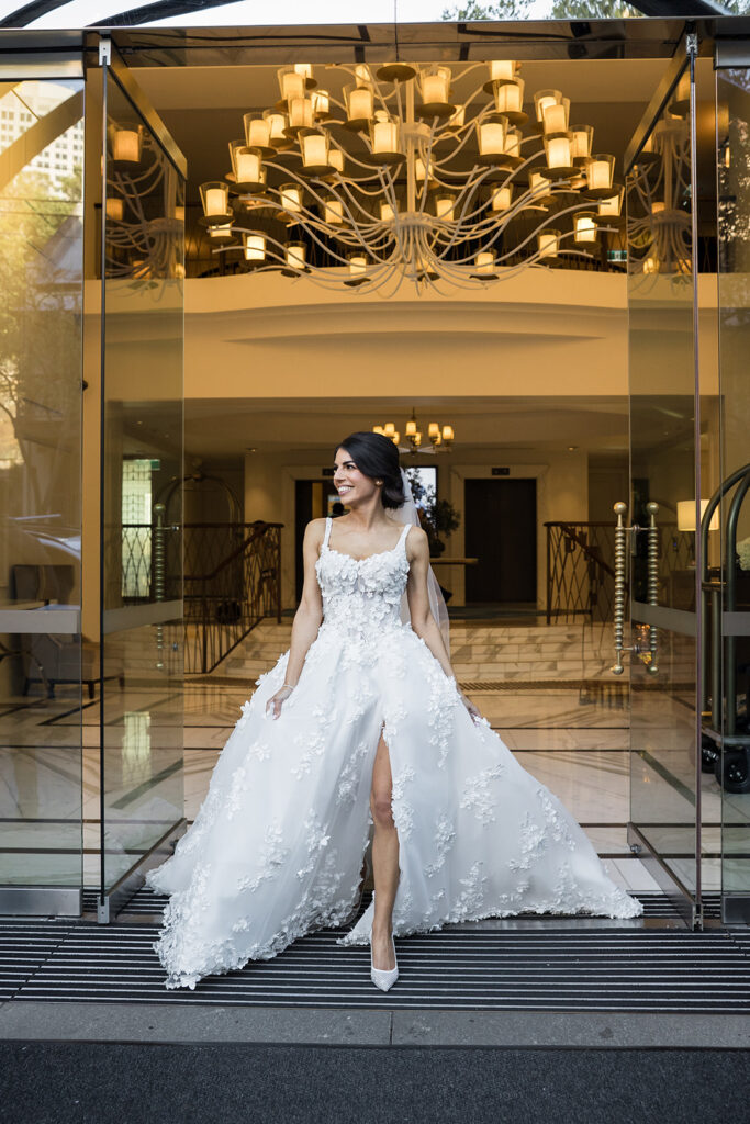 Bride in a white lace wedding gown with floral appliqués and a long train exits a glass doorway, a grand chandelier overhead.