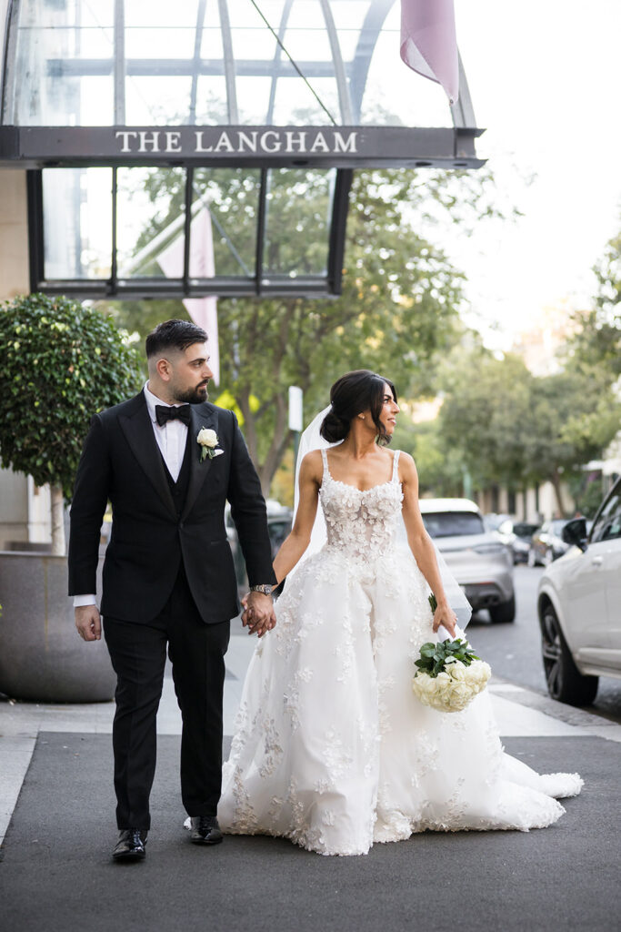 Bride in a white floral-embellished gown holds a bouquet and walks hand in hand with a groom in a black tuxedo down a city street beneath a sign that reads 'THE LANGHAM'.