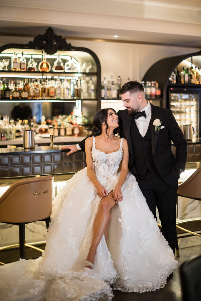 Bride in a lace wedding gown sits at a bar with her groom in a black tux, smiling at each other.