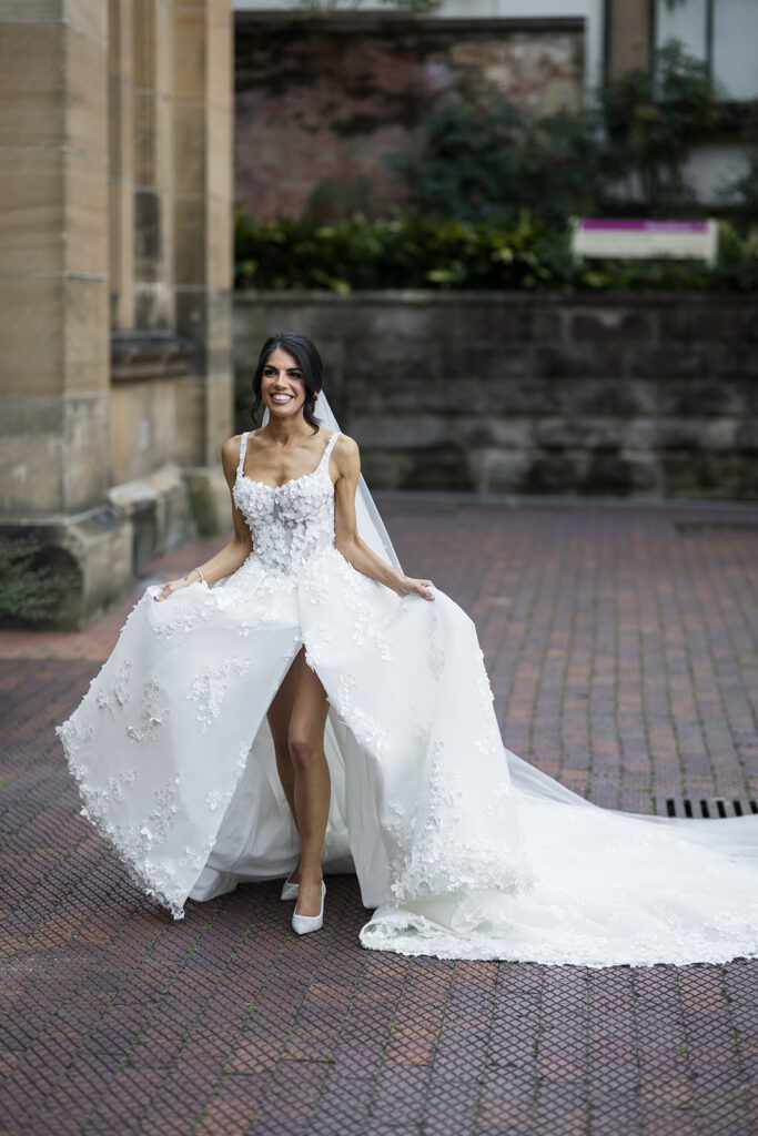 Smiling bride in a white lace wedding gown with a long train standing on a brick-paved walkway.