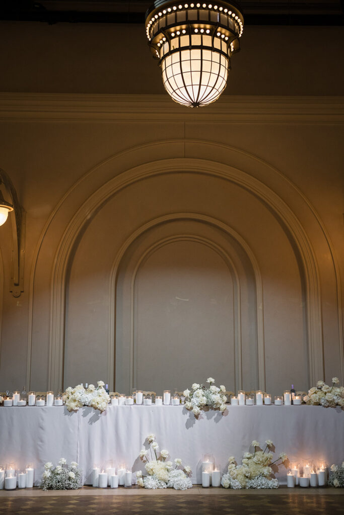 Long banquet table with white tablecloth, candle centers, and white floral arrangements beneath a large arched wall panel and warm chandelier lighting.