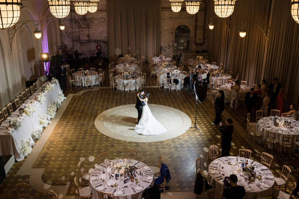 Couple in wedding gown and suit dancing in a circular spotlight at a formal reception hall with round tables nearby and guests watching