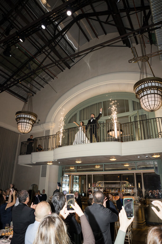 Bride and groom stand on a balcony at a wedding reception, sparks fly from decorative fountains as guests cheer below, many raising phones to capture the moment.