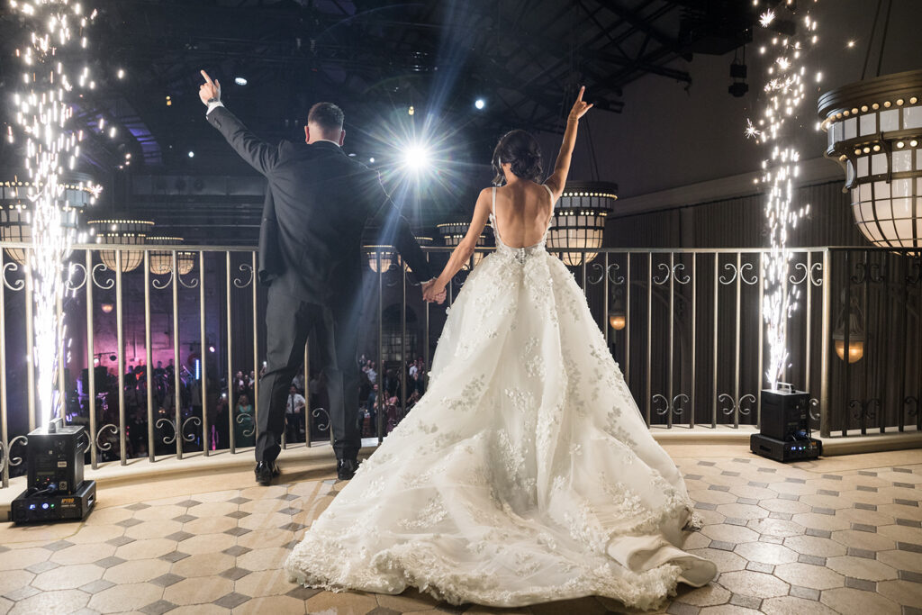 Bride in a voluminous white lace wedding gown and a man in a suit hold hands on a balcony as fireworks erupt nearby.