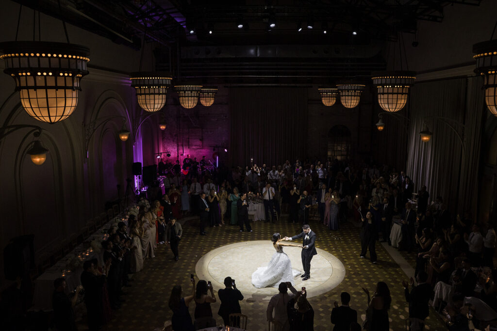 Bride and groom dancing on a circular illuminated dance floor, surrounded by formally dressed guests in a grand ballroom.