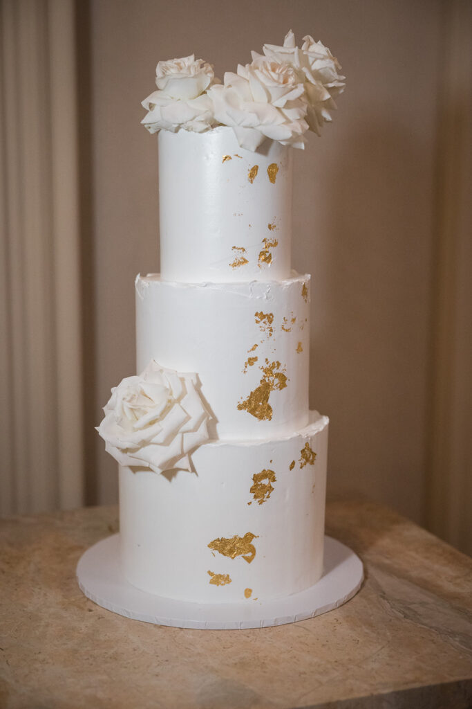 White four-tier wedding cake with gold leaf flecks and white roses on top and side, on a cake stand.