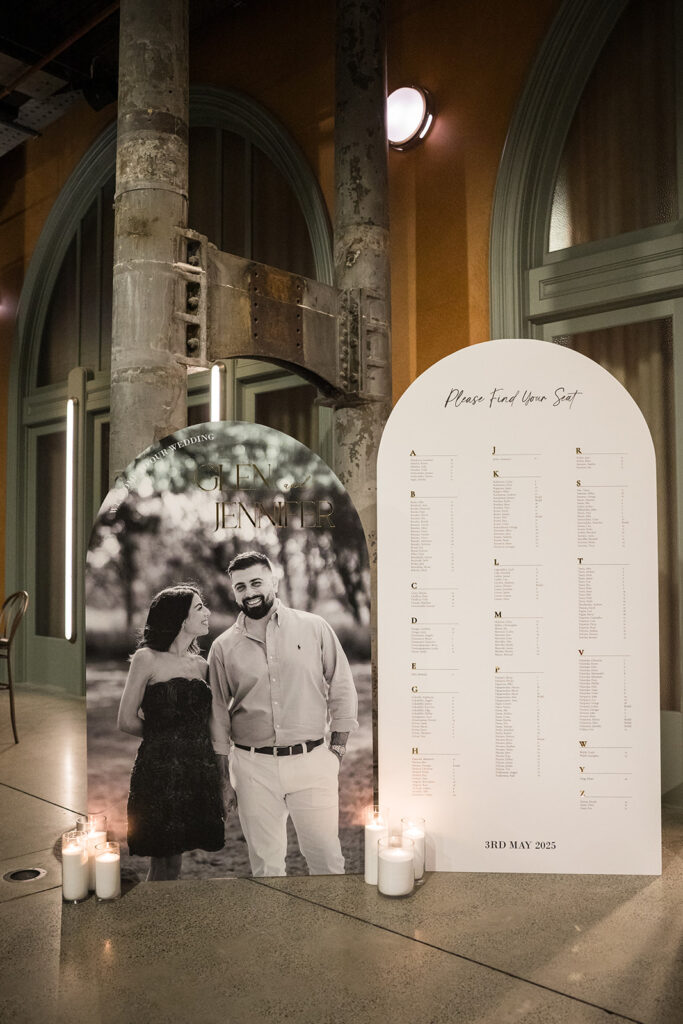 Wedding seating chart board with 'Please find your seat' next to a couple's portrait and lit candles at a venue entrance.