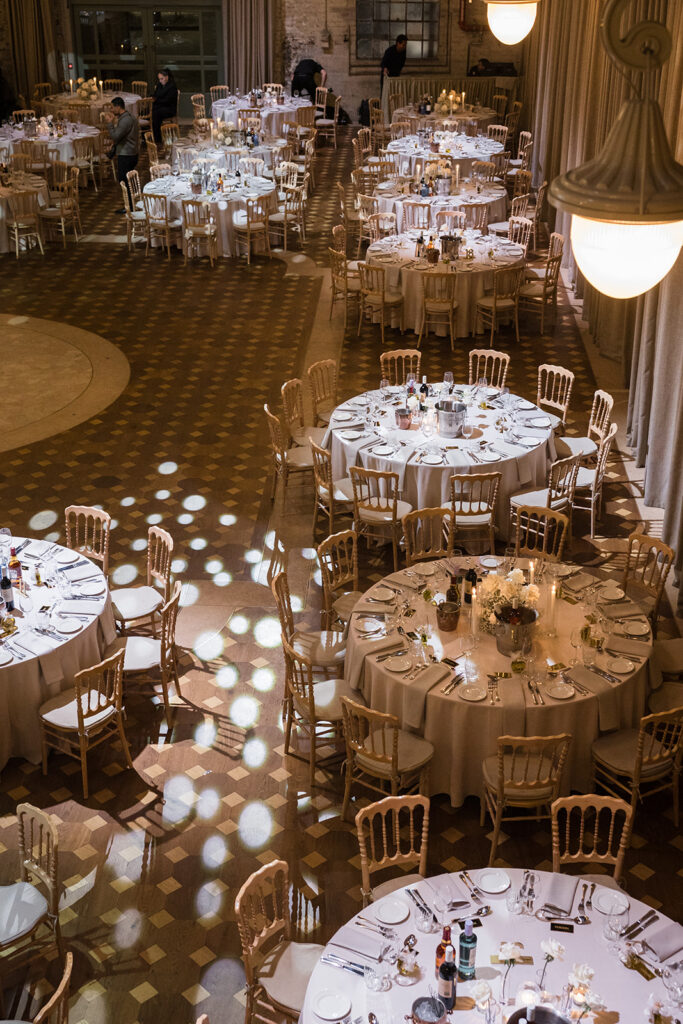 Banquet hall with round tables in white tablecloths, set with silverware, glassware, candles, and cream chairs under warm lighting.