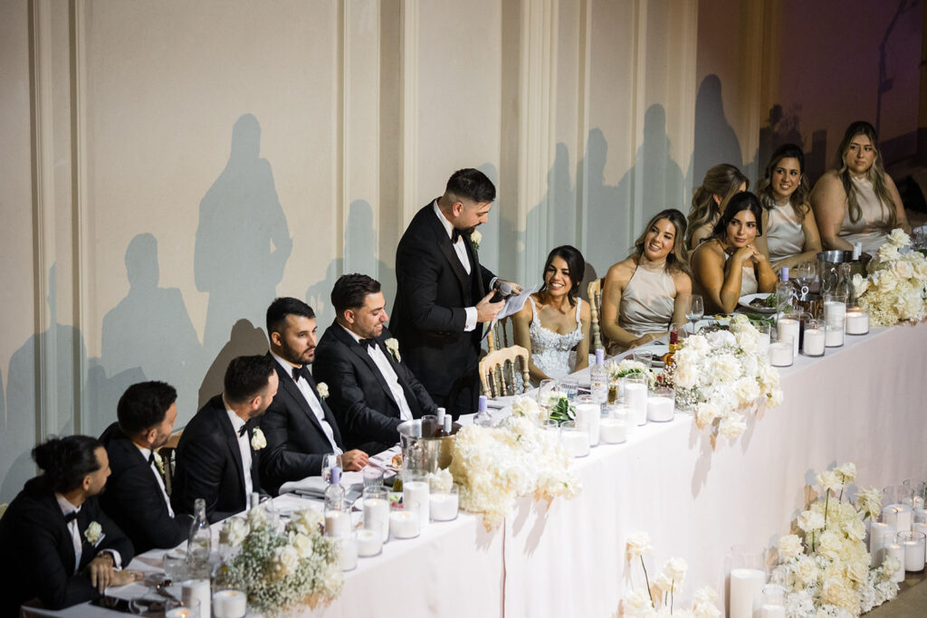 Groom giving a speech at the head table with bridesmaids smiling nearby during a wedding reception.