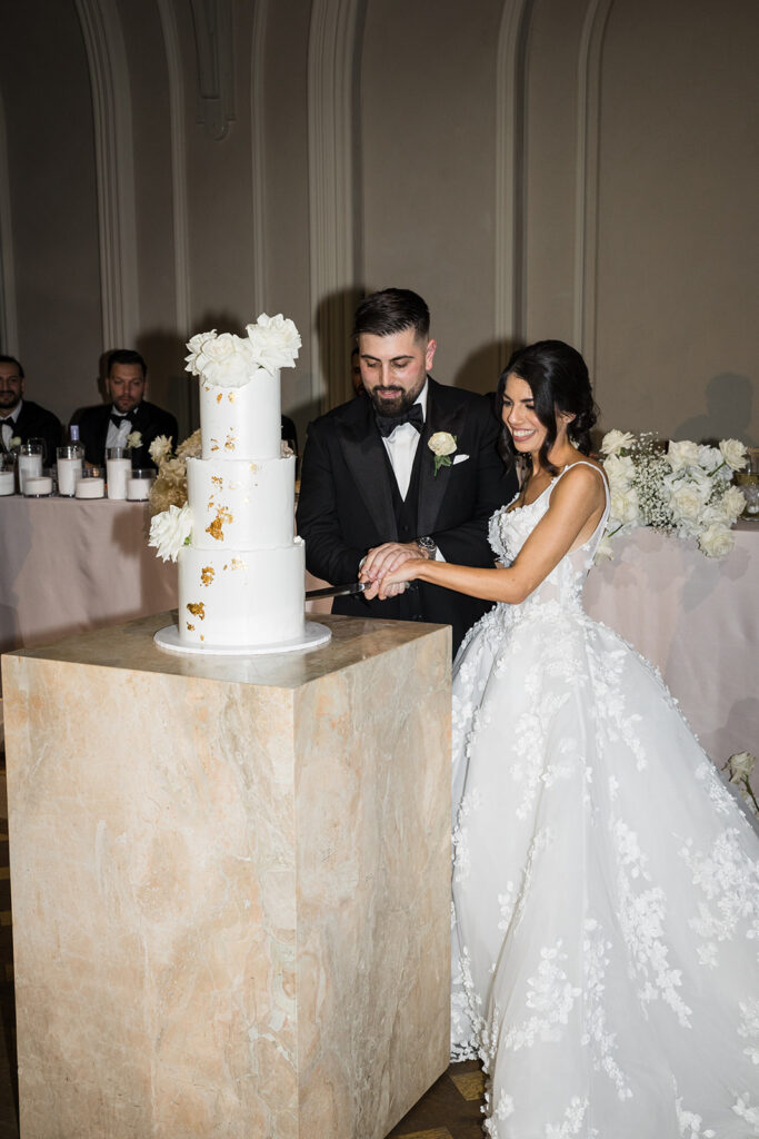 Bride and groom in tuxedo and lace gown cut a tall white wedding cake with white flowers on a marble pedestal.",