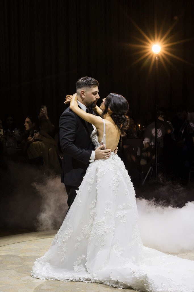Bride and groom share a close dance during their wedding reception, the bride in a lacey white gown and the groom in a dark suit, surrounded by stage fog and a warm spotlight.