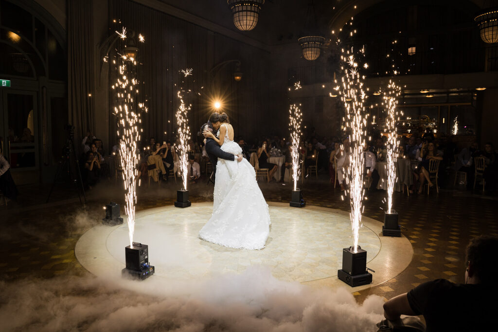 Bride and groom in a white wedding gown and dark suit share a kiss while circling on a lit circular dance floor with fireworks.