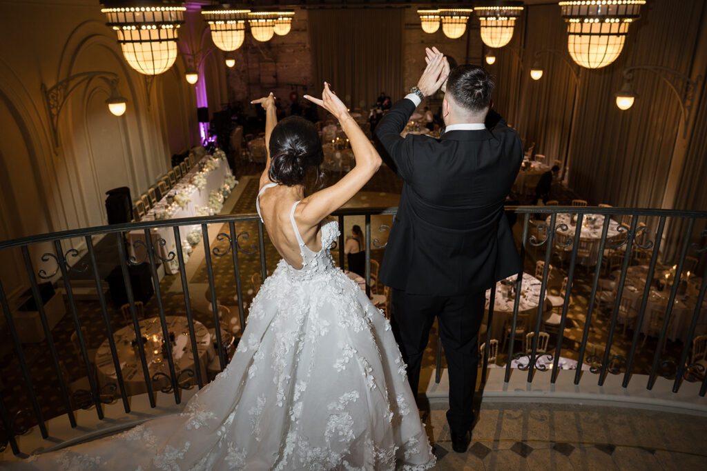 Bride in a white floral wedding gown and a groom in a black suit raise their arms on a balcony overlooking a decorated banquet hall below.